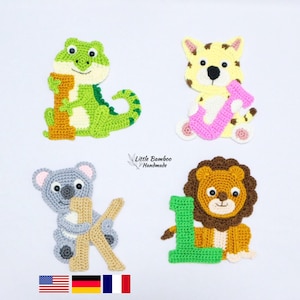 Puede incluir: Cuatro apliques de animales de ganchillo: un caimán verde, un tigre amarillo, un koala gris y un león marrón. Cada animal sostiene el número "1" en un color diferente. También se ve el texto "Little Bamboo Handmade".