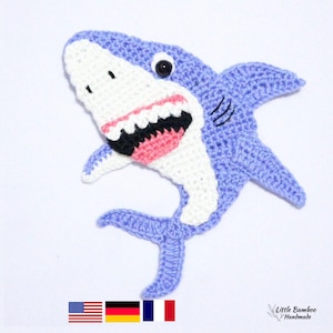 May include: A handmade crocheted shark applique in shades of blue, white, black, and pink. The shark has an open mouth with visible teeth and a single black eye. The applique is handmade.