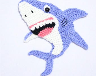 May include: A crocheted shark applique in shades of blue, white, black, and pink. The shark has a wide open mouth with visible teeth and a single black eye. The applique is handmade.