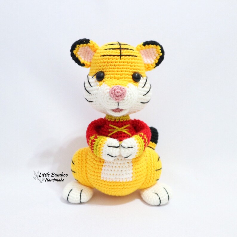 May include: A handmade crochet tiger doll in yellow, black, white, and red. The tiger has a yellow body, black stripes, and a red shirt. The doll is sitting with its paws together. The text "Little Bamboo Handmade" is in the lower left corner.