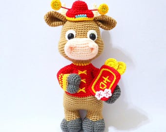 May include: A handmade crochet ox doll in tan, red, and yellow. The ox wears a red outfit and a red hat with yellow accents. It holds a red packet with Chinese characters and yellow coins. The doll has gray hooves and a light pink muzzle.