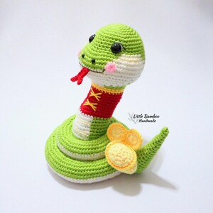 May include: A green, white and red crocheted snake toy with black eyes and a yellow crocheted tail. The snake is wearing a red crocheted collar with white stitching. The text "Little Bamboo Handmade" is visible in the background.