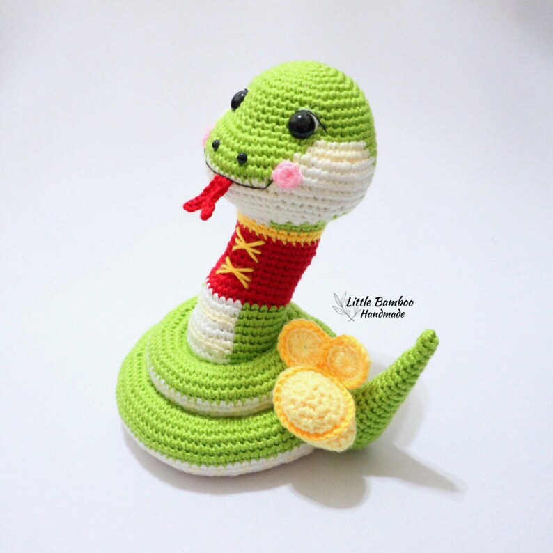 May include: A green, white and red crocheted snake toy with black eyes and a yellow crocheted tail. The snake is wearing a red crocheted collar with white stitching. The text "Little Bamboo Handmade" is visible in the background.