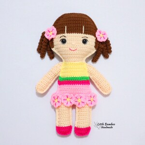 May include: A crocheted doll with brown hair, pink and yellow clothing, and pink flowers on her head and skirt. The doll is standing on a white background.