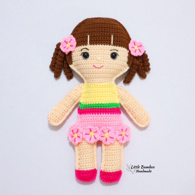 May include: A crocheted doll with brown hair, pink and yellow clothing, and pink flowers on her head and skirt. The doll is standing on a white background.