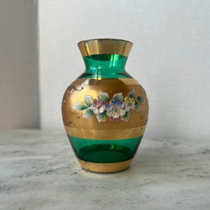 May include: A small green glass vase with gold accents and painted floral details. The vase has a round body and a narrow neck.