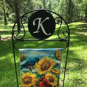 May include: Black metal garden flag stand with a round sign featuring the letter "K" in white. A colorful garden flag with sunflowers, butterflies, and bluebirds is displayed on the stand.