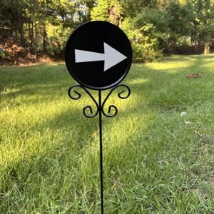 May include: A black circular metal sign featuring a white arrow pointing to the right. The sign is mounted on a thin black metal stake with decorative scrollwork details located directly beneath the circular sign, designed for outdoor placement.