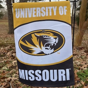 May include: A rectangular garden flag featuring the words "UNIVERSITY OF MISSOURI" in white text on a black and gold background. The flag displays the university's tiger mascot in a black and gold oval.