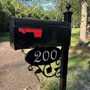 May include: A black metal mailbox mounted on a post, featuring a bright red signal flag on the side. Below the mailbox, an oval black plaque displays the white house number 200, supported by a decorative black metal scrollwork bracket.
