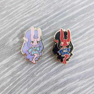 May include: Two enamel pins featuring cartoon characters with bunny and devil ears. The character on the left has purple hair, pink skin, and a blue dress. The character on the right has black hair, red skin, and a blue dress.