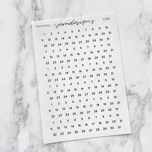 May include: A white sticker sheet with black numbers for dates, arranged in rows and columns. The text "4.5MM DATES" and "jemdesigns" is printed on the top left. The sticker sheet is on a white and grey marble surface.