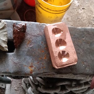 May include: An assortment of handcrafted items, including a copper mold with three circular cavities, two stone arrowheads, and a glass knife with a wooden handle. The items are displayed on a weathered metal surface.
