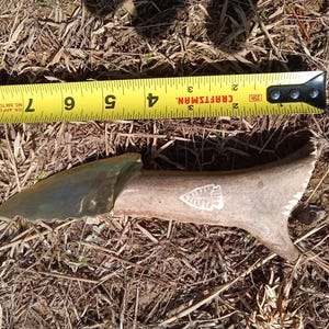 May include: A handmade knife with a green blade and a bone handle. The handle has a white design of a stylized arrowhead. The knife is measured against a yellow tape measure.
