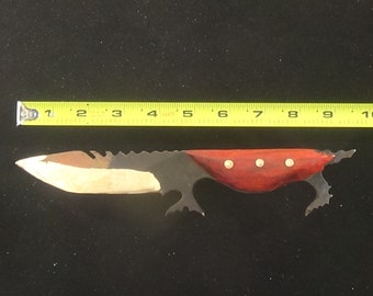 May include: A handmade knife with a black and white blade and a red wooden handle. The knife is approximately 7 inches long and has a serrated edge. The handle has three brass rivets.