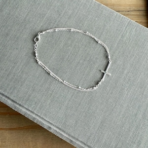 May include: A silver-colored bracelet with a cross charm. The bracelet features a double-strand design with small, round beads and a delicate chain. The cross is positioned on the side of the bracelet.