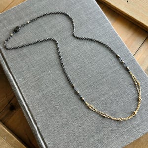 May include: A delicate layered necklace with a black chain and a gold chain, accented with small gold beads.