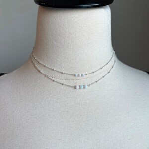 May include: A silver layered necklace with three strands of chain and a row of small white opal beads.