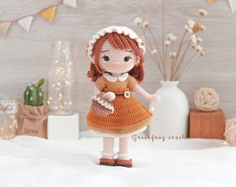 May include: A crocheted doll with brown hair, wearing a white dress with a brown belt and holding a brown crocheted basket. The doll is standing on a white surface with a brown background.
