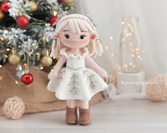 May include: A crocheted doll wearing a white dress with silver accents and brown boots. The doll has blonde hair and a crown. The doll is standing in front of a Christmas tree with red ornaments.
