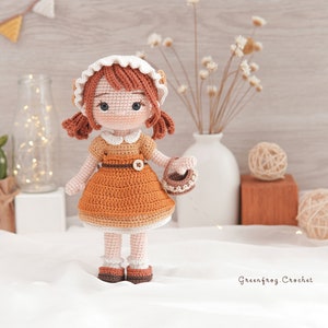 May include: A crocheted doll with brown hair, wearing a white and brown dress and a white bonnet, holding a small basket. The doll is standing on a white surface with a wooden background.