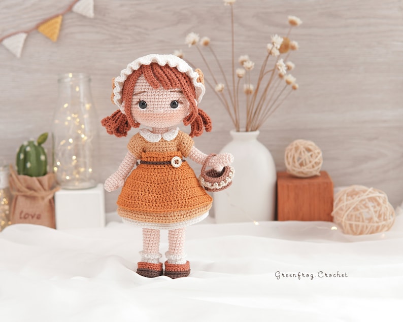 May include: A crocheted doll with brown hair, wearing a white and brown dress and a white bonnet, holding a small basket. The doll is standing on a white surface with a wooden background.