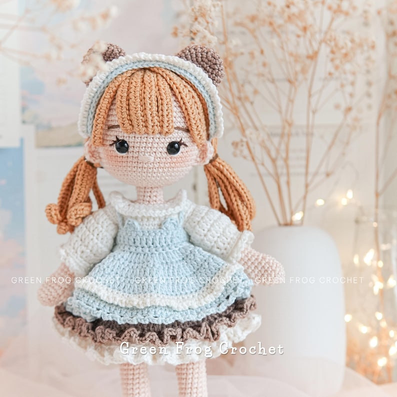 Peut inclure: Une poup&eacute;e en crochet avec des cheveux bruns, une robe bleue avec une bordure en dentelle blanche et des oreilles en crochet brun. La poup&eacute;e porte une chemise en crochet blanc et a des yeux noirs. La poup&eacute;e est debout sur une surface blanche avec un vase de fleurs s&eacute;ch&eacute;es en arri&egrave;re-plan.