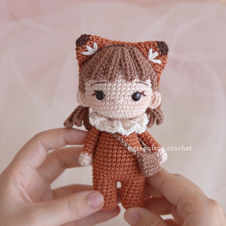 May include: A crocheted doll wearing a fox costume with brown and white fur accents. The doll has brown hair, a white face, and brown eyes. The doll is holding a small brown bag. The doll is wearing a brown crocheted outfit with a white crocheted collar. The doll is standing on a person's hand. The text "@greenfrog.crochet" is visible in the image.