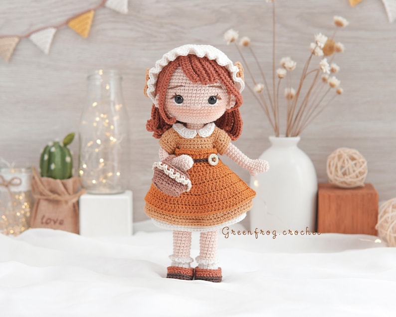 May include: A crocheted doll with brown hair, wearing a white dress with a brown belt and holding a brown crocheted basket. The doll is standing on a white surface with a brown background.