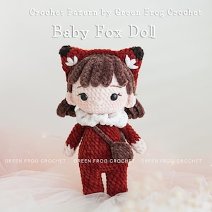 May include: Crocheted baby fox doll wearing a red and brown outfit with a white collar and a brown satchel. The doll has brown hair and a pink face with black eyes. The doll is sitting on a white surface with white beads.