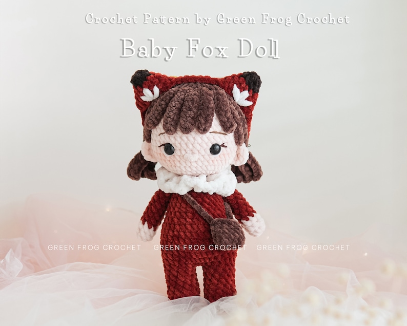 May include: Crocheted baby fox doll wearing a red and brown outfit with a white collar and a brown satchel. The doll has brown hair and a pink face with black eyes. The doll is sitting on a white surface with white beads.