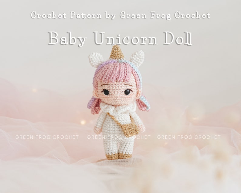 Puede incluir: Una mu&ntilde;eca de beb&eacute; unicornio de crochet con pelo rosa, un cuerno blanco y un atuendo blanco y marr&oacute;n. La mu&ntilde;eca est&aacute; sentada sobre un fondo de tela rosa y blanca.