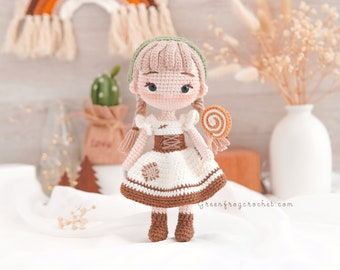 May include: A crocheted doll with brown hair, a green headband, and a white dress with brown accents. The doll is holding a swirl lollipop and is standing on a white surface.