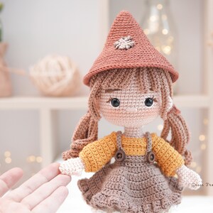 May include: A crocheted doll with brown hair, wearing a brown and orange dress, a brown hat with a flower detail, and brown shoes. The doll is holding out its hand.