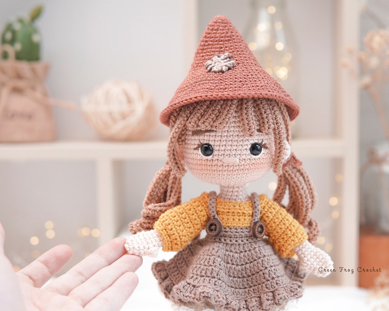 May include: A crocheted doll with brown hair, wearing a brown and orange dress, a brown hat with a flower detail, and brown shoes. The doll is holding out its hand.