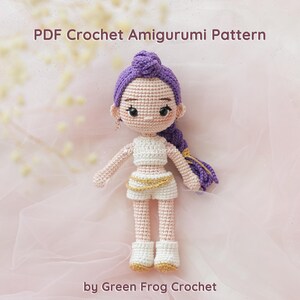 May include: A crocheted amigurumi doll with purple hair styled in a bun and braid. The doll wears a white crop top, shorts, and boots with gold accents. The text "PDF Crochet Amigurumi Pattern" and "by Green Frog Crochet" are visible.