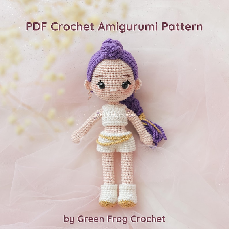 May include: A crocheted amigurumi doll with purple hair styled in a bun and braid. The doll wears a white crop top, shorts, and boots with gold accents. The text "PDF Crochet Amigurumi Pattern" and "by Green Frog Crochet" are visible.