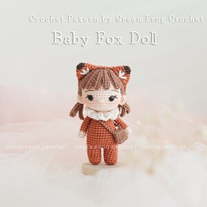 May include: Crocheted amigurumi doll of a fox wearing a brown and white outfit with a small brown bag. The doll has brown hair, a white face, and black eyes. The doll is standing on a wooden base. The text "Crochet Pattern by Green Frog Crochet" and "Baby Fox Doll" is above the doll.
