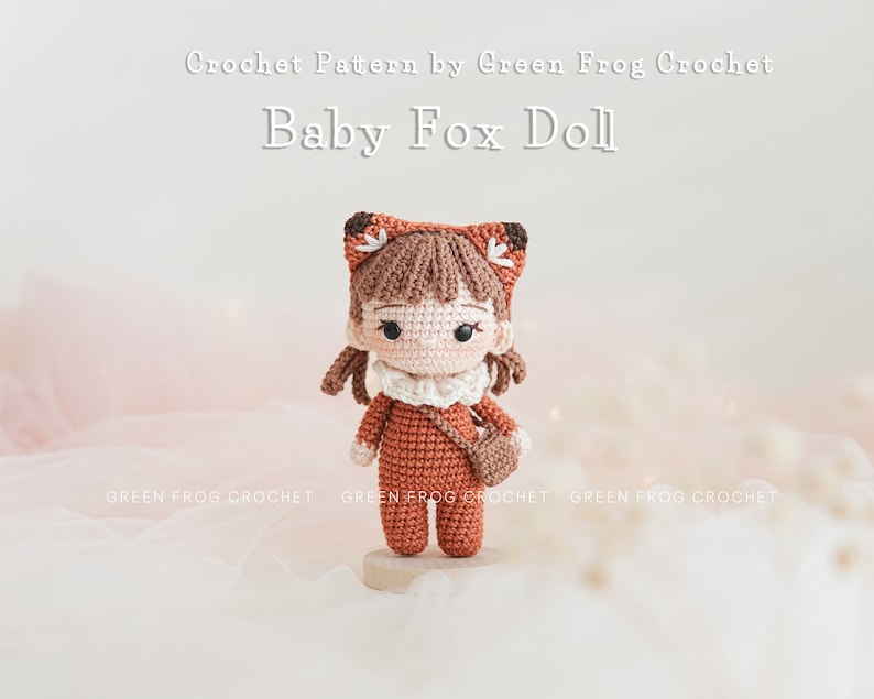 May include: Crocheted amigurumi doll of a fox wearing a brown and white outfit with a small brown bag. The doll has brown hair, a white face, and black eyes. The doll is standing on a wooden base. The text "Crochet Pattern by Green Frog Crochet" and "Baby Fox Doll" is above the doll.