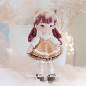 Peut inclure: Une poup&eacute;e en crochet avec des cheveux bruns, une robe blanche et marron et des chaussures marrons. La poup&eacute;e est debout sur une surface blanche.