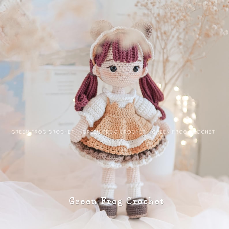 Peut inclure: Une poup&eacute;e en crochet avec des cheveux bruns, une robe blanche et marron et des chaussures marrons. La poup&eacute;e est debout sur une surface blanche.