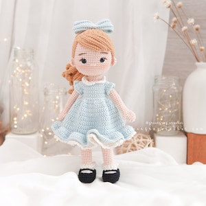 Puede incluir: Una mu&ntilde;eca de crochet con cabello casta&ntilde;o, un vestido azul y zapatos negros. La mu&ntilde;eca lleva un lazo azul en el pelo.