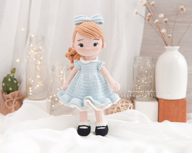 Puede incluir: Una mu&ntilde;eca de crochet con cabello casta&ntilde;o, un vestido azul y zapatos negros. La mu&ntilde;eca lleva un lazo azul en el pelo.