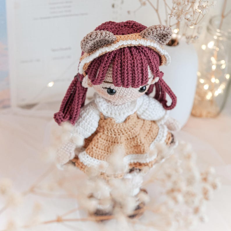 Peut inclure: Une poup&eacute;e en crochet avec des oreilles brunes et blanches, un chapeau bordeaux et une robe bordeaux et brune. La poup&eacute;e a de longs cheveux bordeaux et des yeux bruns.