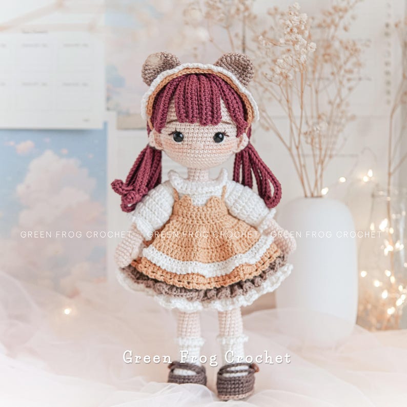 Peut inclure: Une poup&eacute;e en crochet avec des oreilles d'ours brun, une robe blanche avec des accents bruns et beiges, et des chaussures brunes. La poup&eacute;e a de longs cheveux roux fonc&eacute;s et un visage brun clair avec des yeux bruns. La poup&eacute;e est debout sur une surface blanche avec un fond blanc.