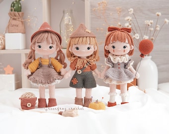May include: Three crocheted dolls with different outfits and accessories. The doll on the left is wearing a brown dress with a yellow top and a brown hat. The doll in the middle is wearing a brown and green dress with a brown hat. The doll on the right is wearing a gray dress with a brown hat and holding a balloon.