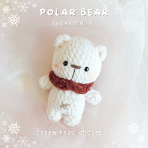 May include: White crocheted polar bear standing with a red scarf. The bear has black eyes and a brown nose. The background is a pink and white fabric with snowflakes.
