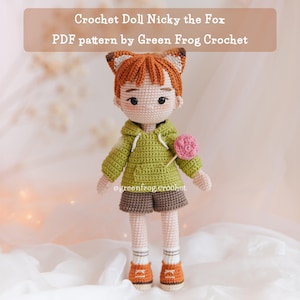May include: A crocheted doll of Nicky the Fox, dressed in a green hoodie, brown shorts, and orange trainers. The doll features fox ears and holds a pink lollipop. The text "Crochet Doll Nicky the Fox" and "PDF pattern by Green Frog Crochet" are visible.