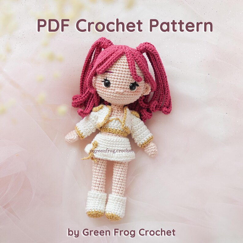May include: A handmade crochet doll with pink hair in pigtails, wearing a white outfit with gold trim. The doll has a light skin tone and is standing. The text "PDF Crochet Pattern" and "by Green Frog Crochet" are visible.