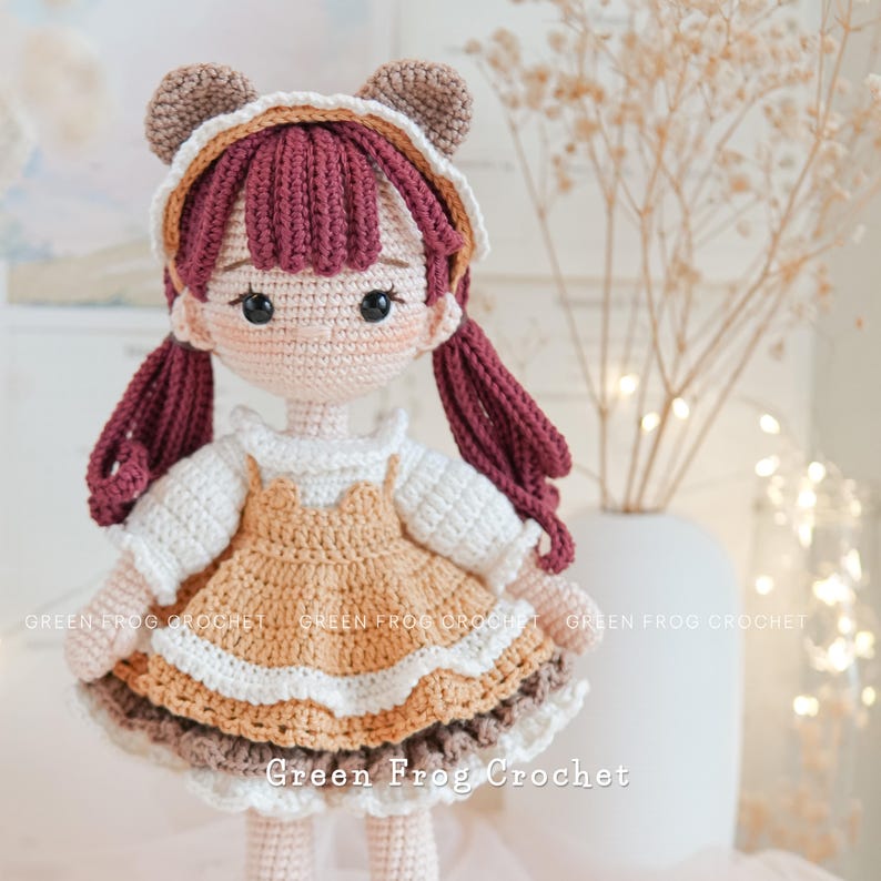 Peut inclure: Poup&eacute;e en crochet avec des oreilles d'ours brun, une robe blanche avec une jupe marron et blanche, et de longs cheveux bruns. La poup&eacute;e est debout sur une surface blanche avec un vase de fleurs s&eacute;ch&eacute;es en arri&egrave;re-plan. Le texte "Green Frog Crochet" est visible en bas de l'image.